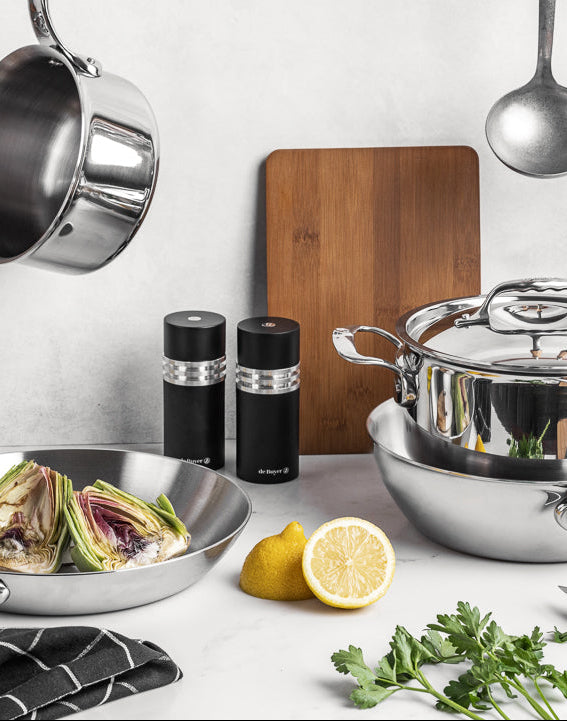 Kitchen counter with stainless steel cookware, lemons, artichokes, and herbs on a white background.