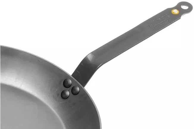 Close-up of a dark gray carbon Steel Mineral B skillet showing the interior rim, rivets, and the long flat handle with a hanging hole and small yellow accent against a white background.